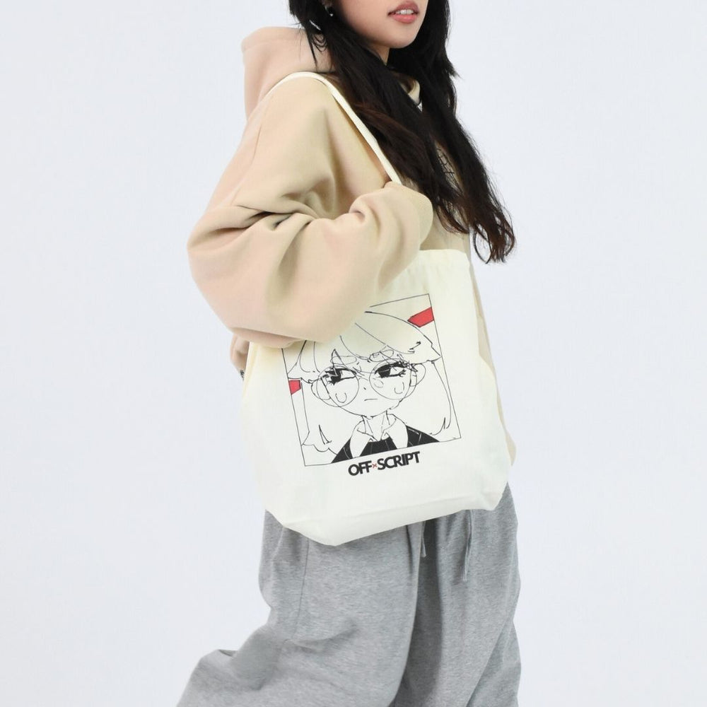 A person wearing a beige sweater and gray pants is holding a white tote bag with a printed character design.